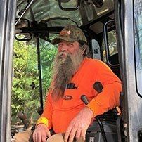 Man with a long beard in an orange shirt and cap, operating heavy machinery.