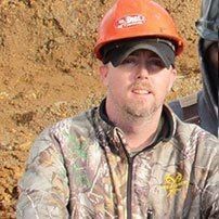 Man wearing an orange hard hat, camouflage jacket, smiling, outdoors.