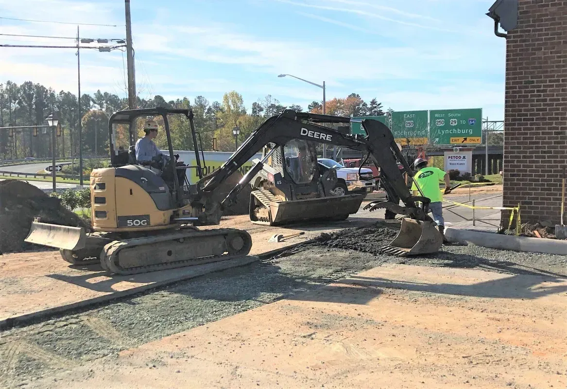 Excavator digging gravel on pavement, with a worker assisting. Sunny day.