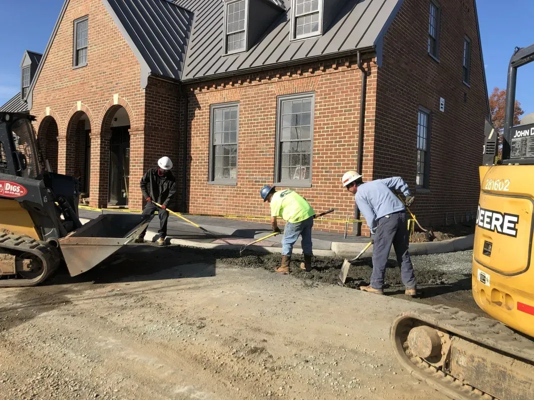 Construction workers paving a brick building's sidewalk with a backhoe and shovels.