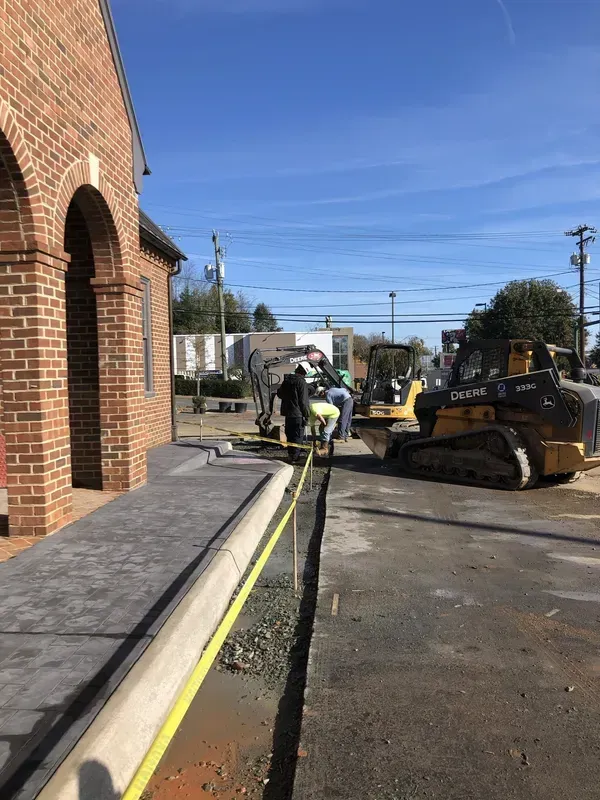 Construction workers with machinery alongside a brick building and a newly poured sidewalk.