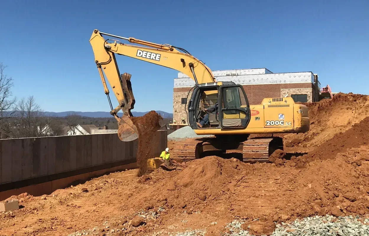 Yellow excavator digging dirt near a building under a clear sky. A worker is visible nearby.