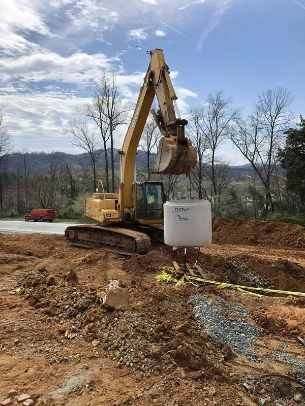 Yellow excavator lifting a cylindrical white object at a construction site, blue sky in background.