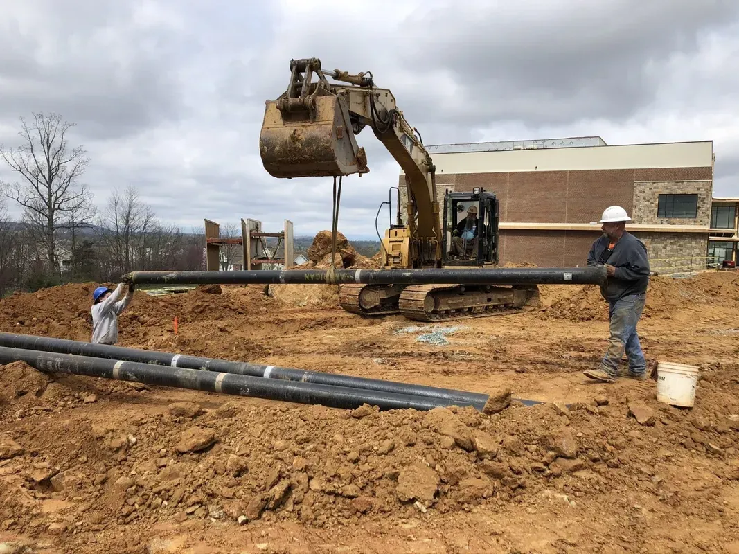 Construction workers installing pipes with an excavator on a dirt construction site. Cloudy sky in the background.
