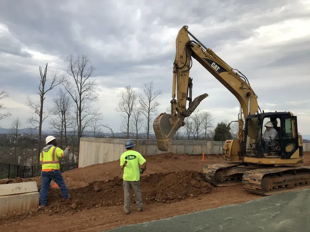 Construction workers operate excavator at a building site. Man in vest directs. Overcast day.