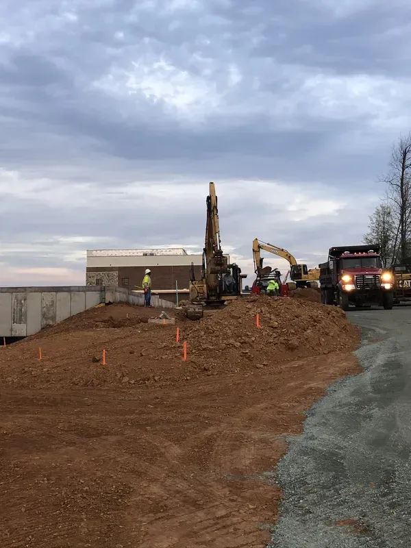 Construction site: Excavator and dump trucks on a dirt hill, workers in vests. Overcast sky.