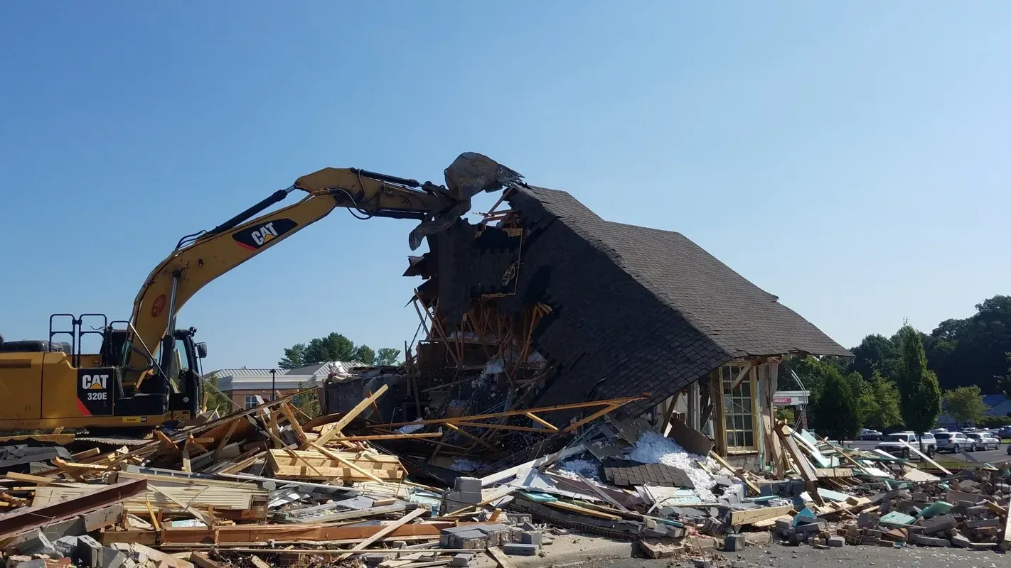 Yellow excavator demolishing a building with a dark shingled roof under a clear blue sky. Debris scattered below.