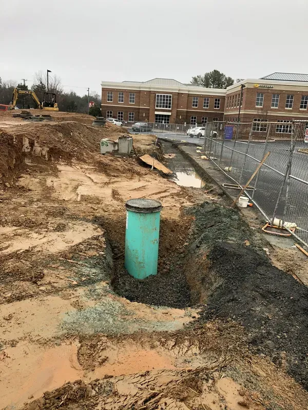 Construction site with a teal pipe in an excavated area, building in background, gray overcast sky.