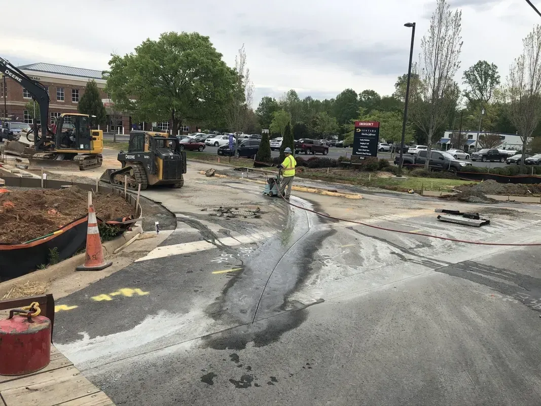 Construction site with heavy machinery and a worker in a yellow vest. Asphalt and dirt are visible.