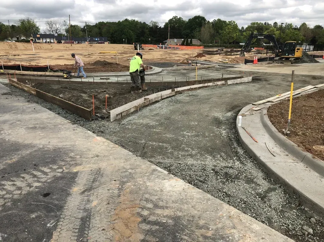 Construction site with workers pouring concrete for a sidewalk, light brown dirt and green trees in the background.
