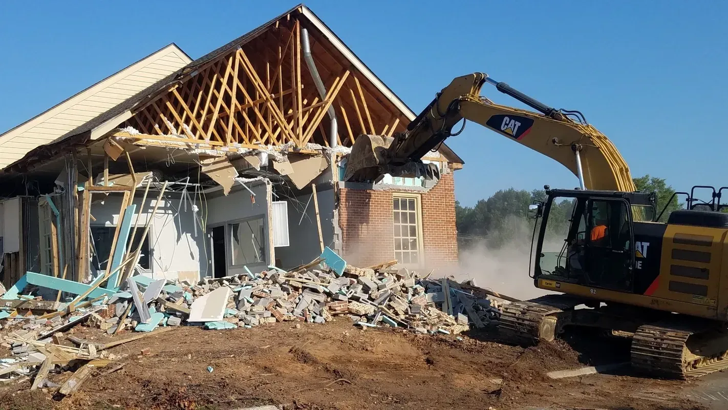 A yellow excavator demolishes a building with wooden beams, brick, and debris under a clear sky.