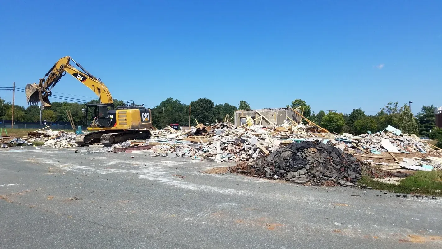 A large excavator demolishes a building on a bright day; piles of debris surround the machine, blue sky.