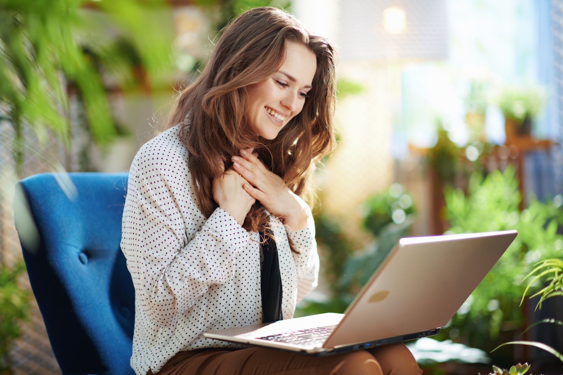 Woman smiling while using a laptop on a blue chair in a bright indoor space