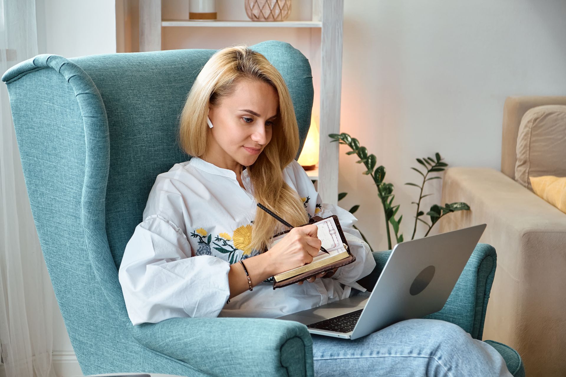 Woman in a teal armchair writing in a notebook beside a laptop in a bright living room
