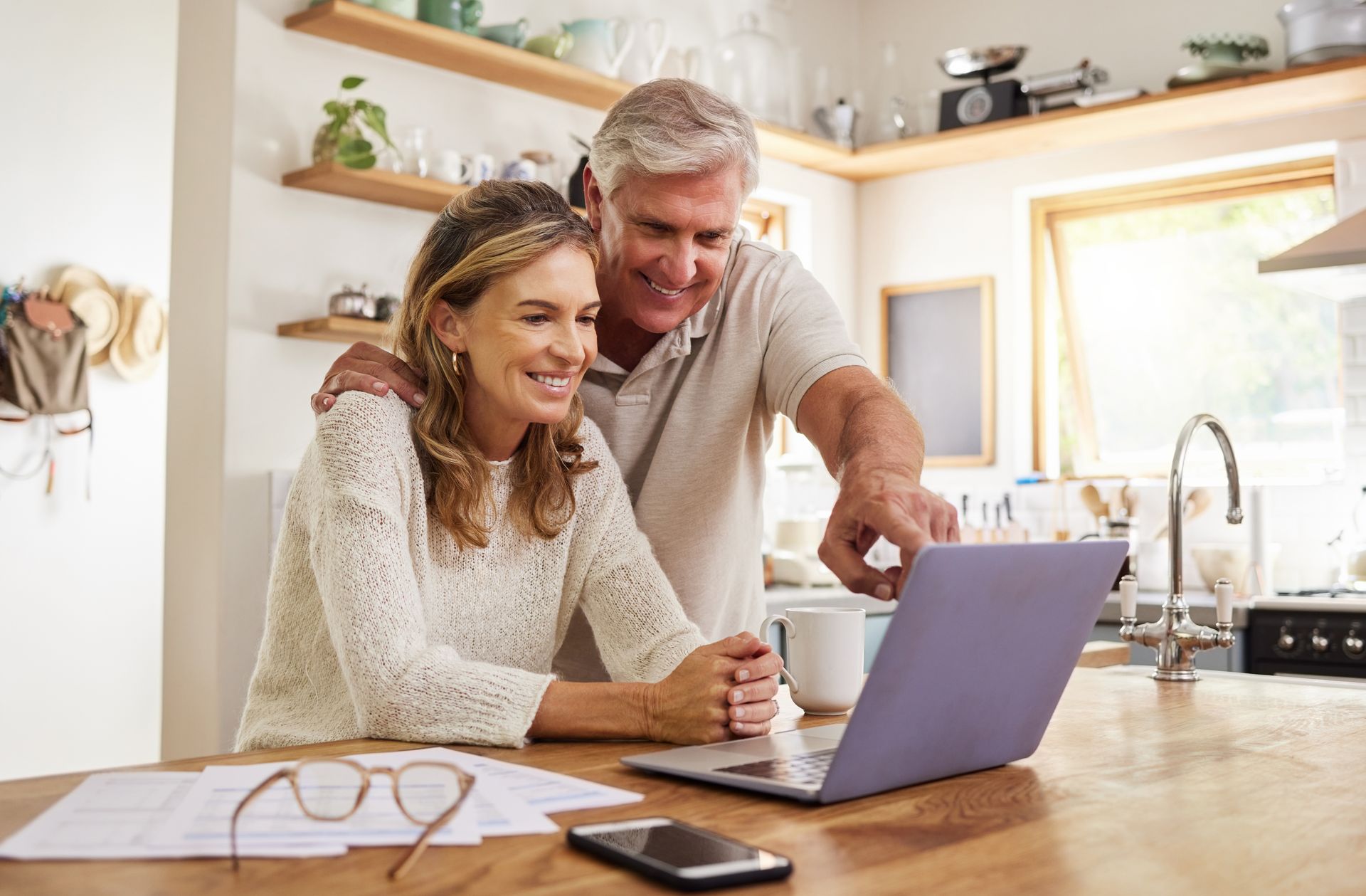 Smiling couple looking at a laptop in a bright kitchen with a coffee mug and glasses on the table