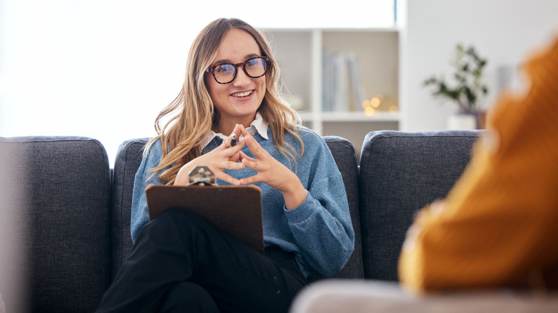 Therapist speaking with a client in a bright office, seated on a gray couch with a clipboard.