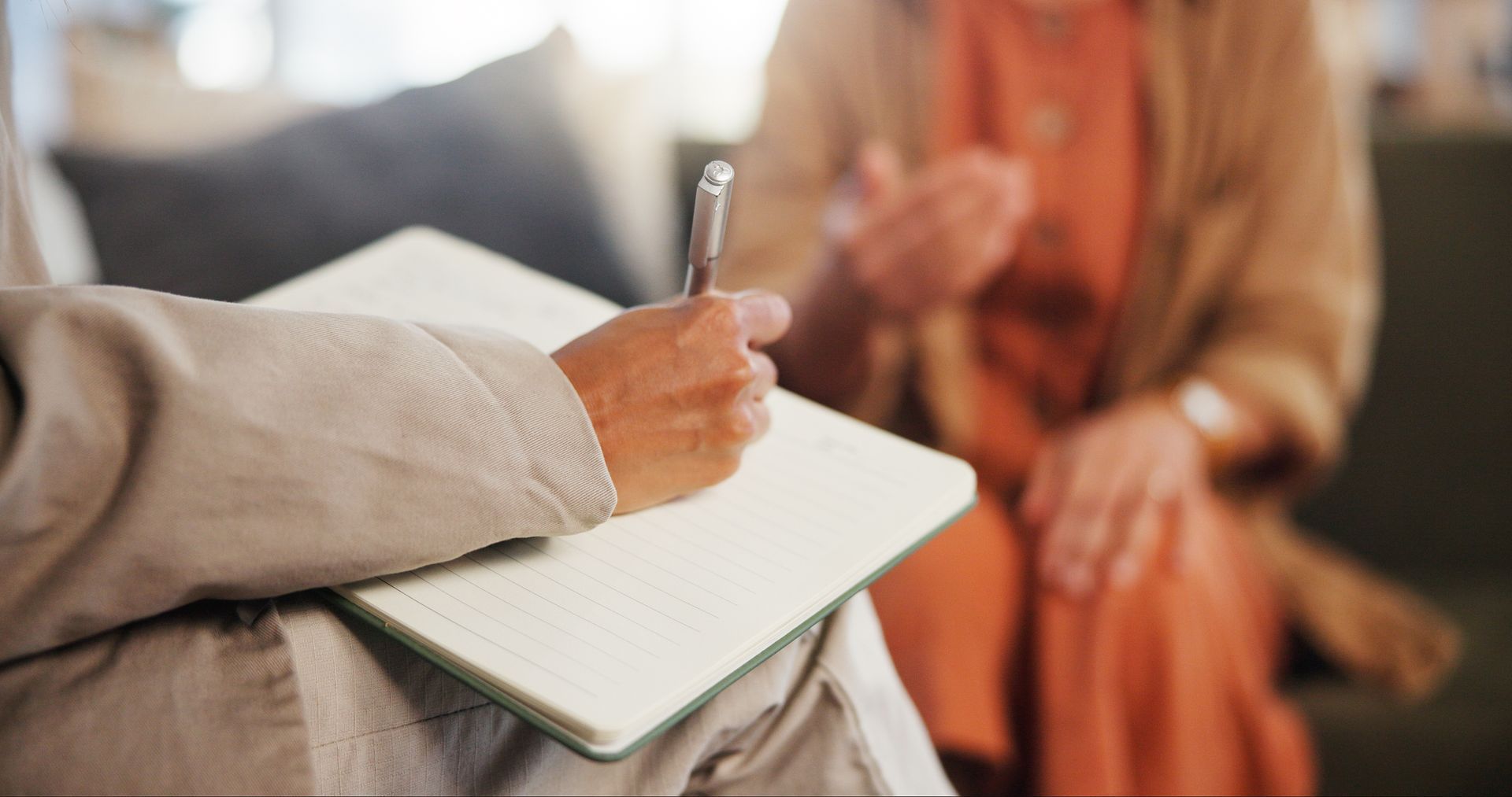 Person writing in a notebook during a conversation with another seated person indoors