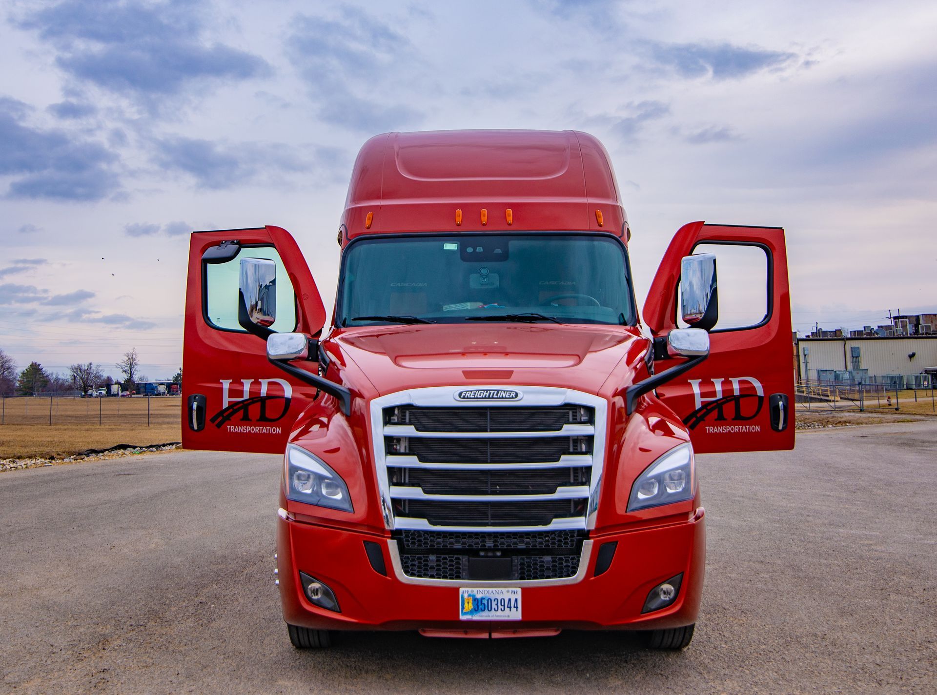 Red Freightliner semi-truck with open doors parked on a gravel road against a cloudy sky.