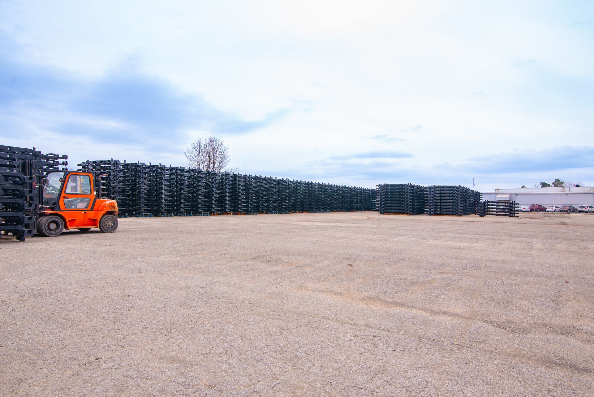 An orange forklift in a gravel lot next to large stacks of black tires under a cloudy sky.