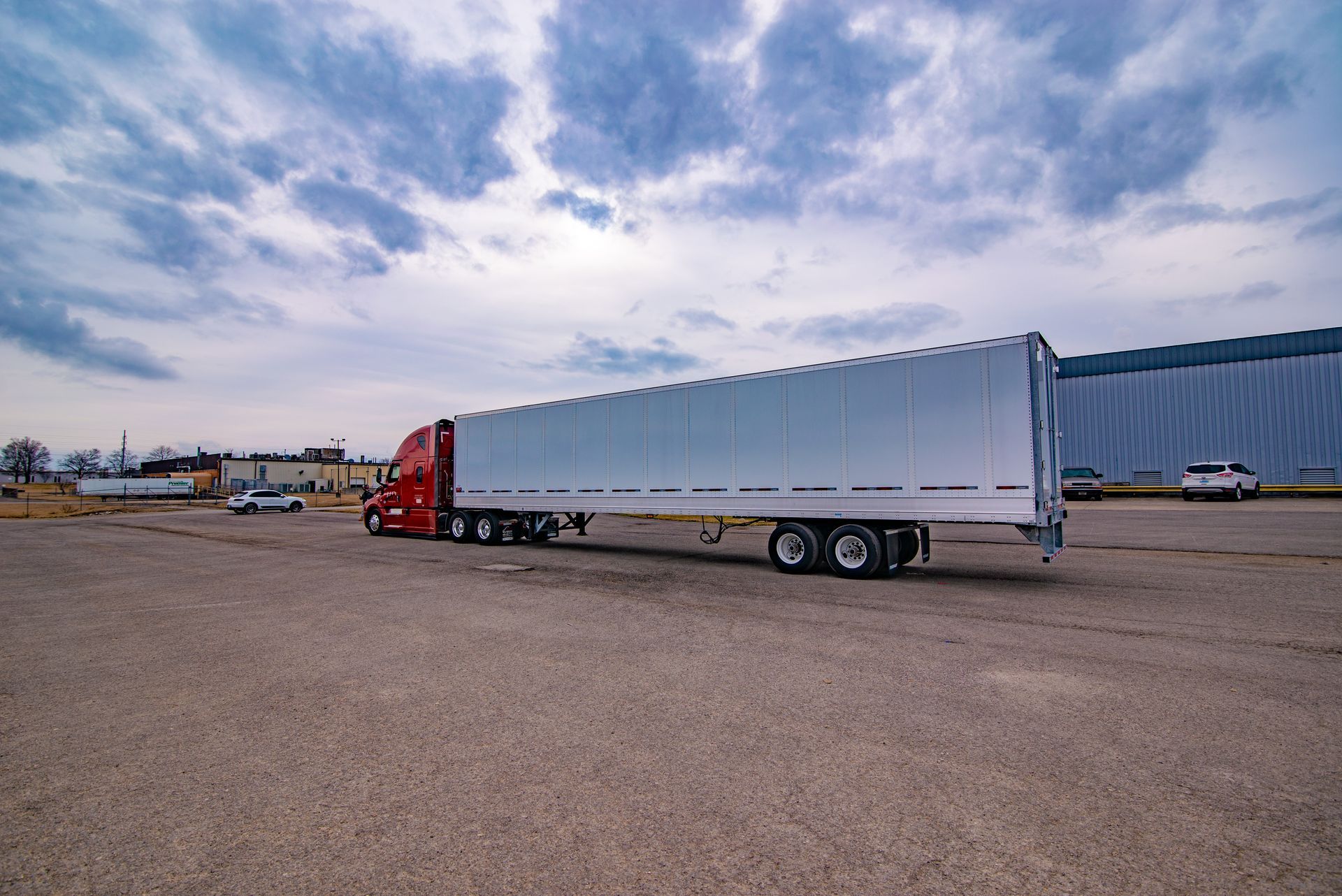Red semi-truck with a long white trailer parked on a gravel lot under a cloudy sky.
