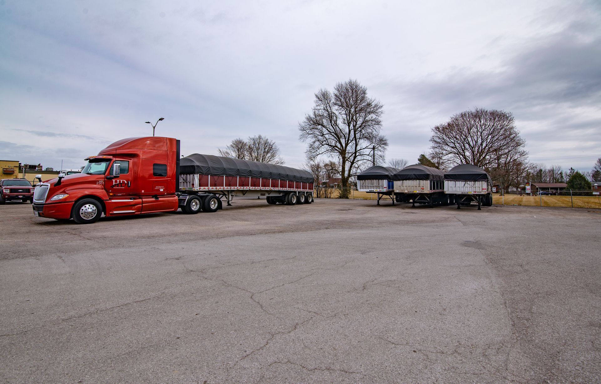 Red semi-truck with two connected trailers parked on gravel lot, overcast sky.