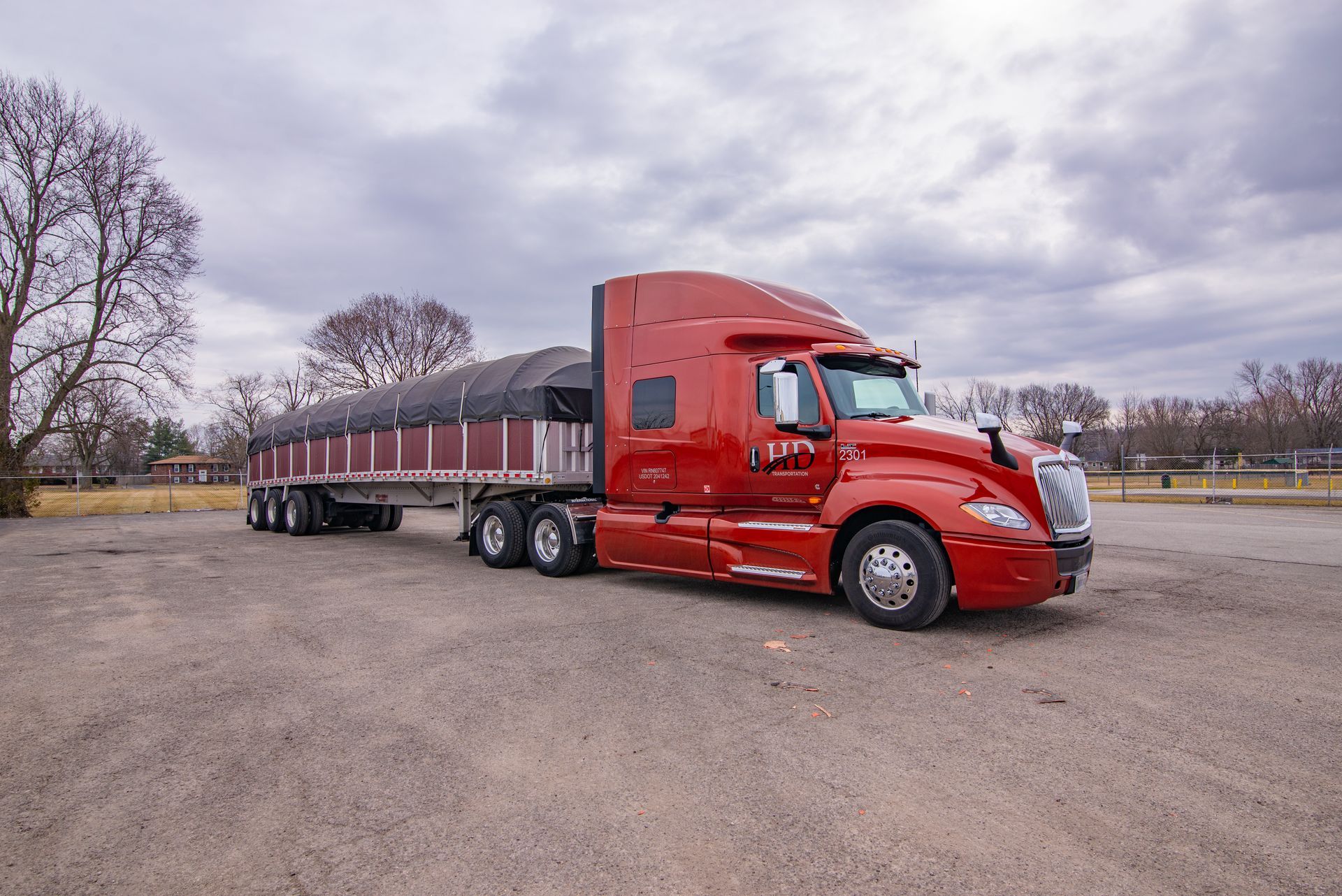 Red semi-truck with a trailer on a gravel lot under a cloudy sky.