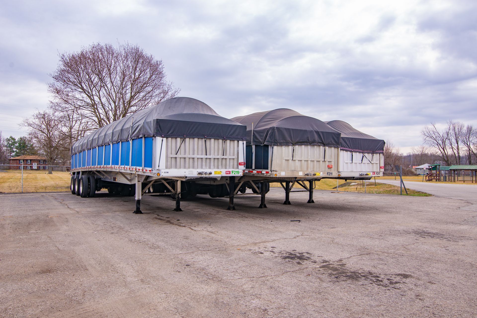 Three parked semi-trailer trucks with black tarps over their contents, on a gravel lot under a cloudy sky.