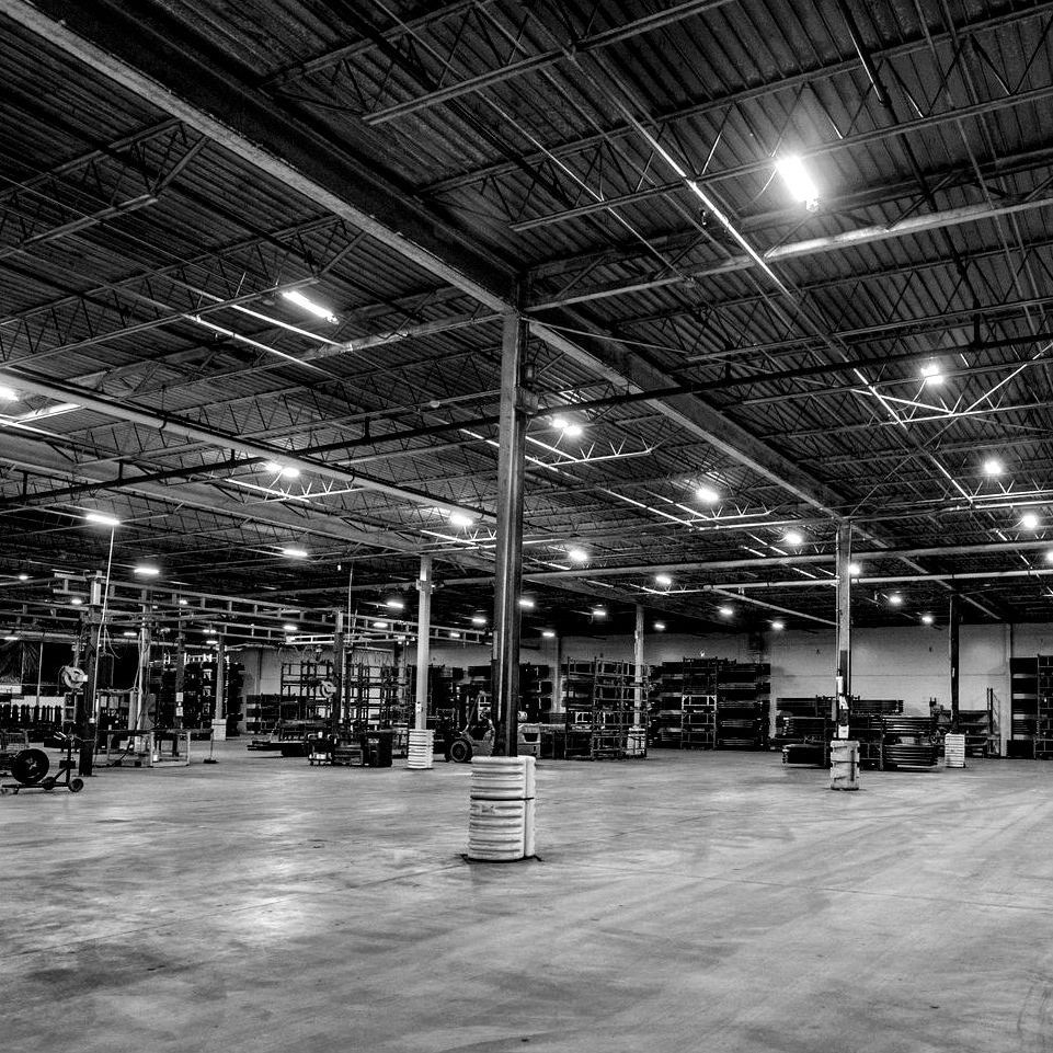 Warehouse interior, black and white. Metal shelving, concrete floor, overhead lighting, support beams.