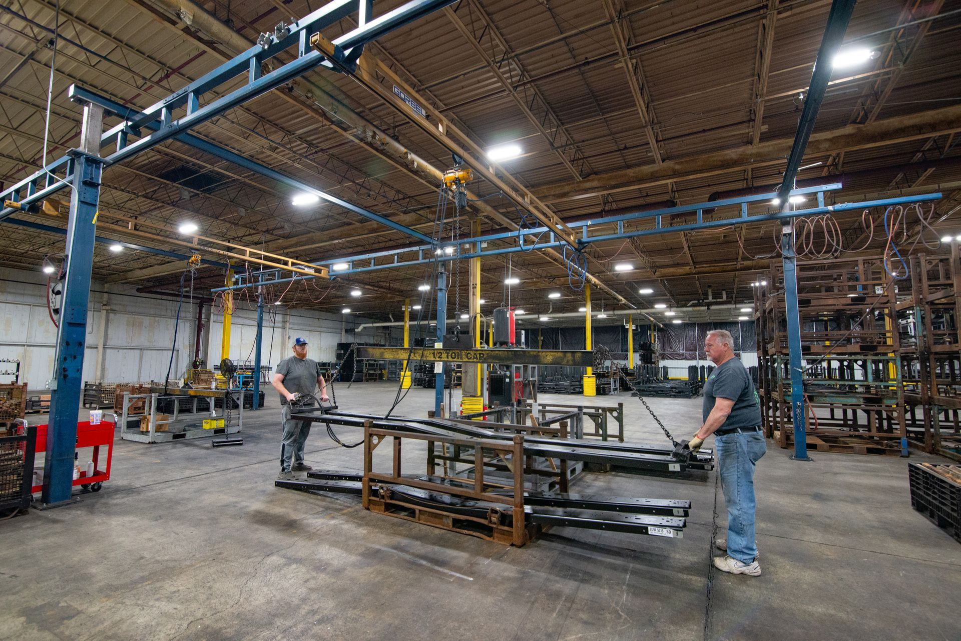 Two workers moving metal bars in a large warehouse with overhead crane system.