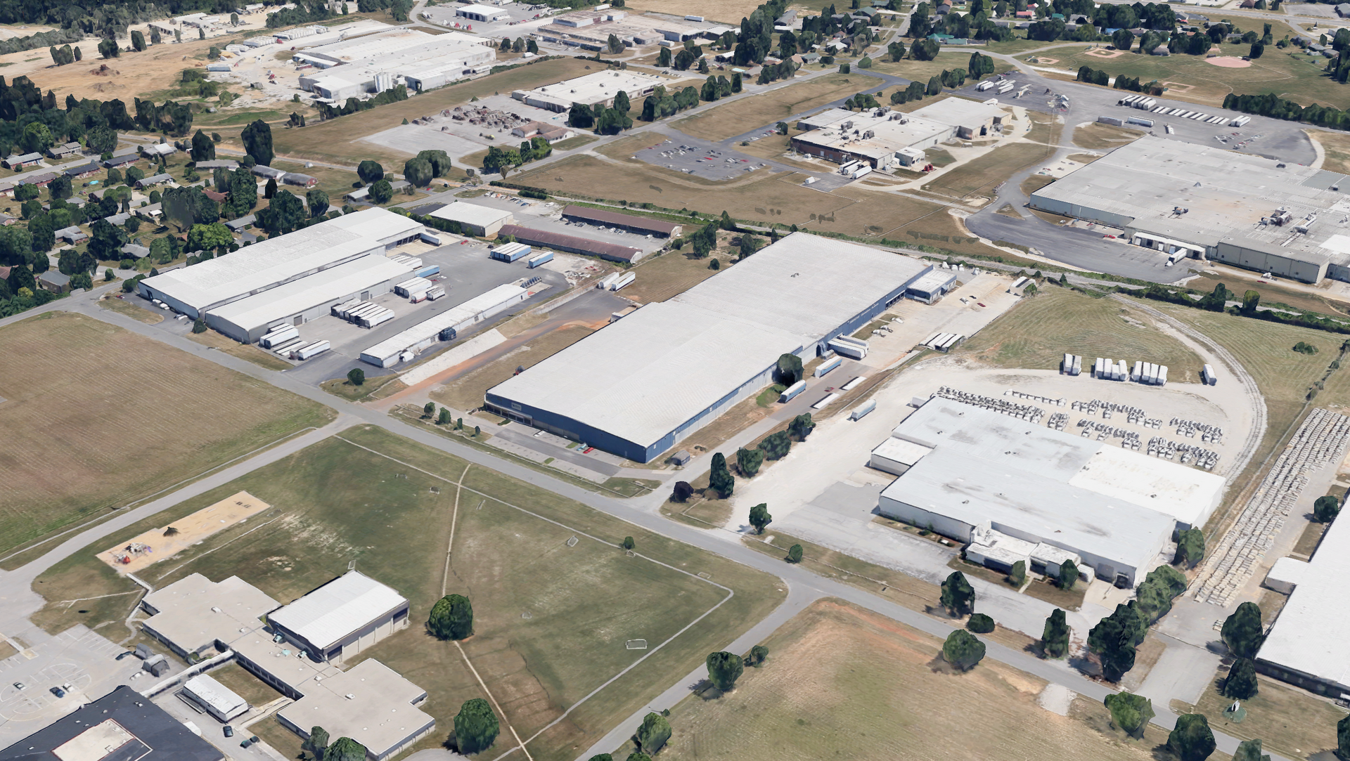 Aerial view of an industrial park with several large warehouses, parking lots, and green spaces under a bright sky.