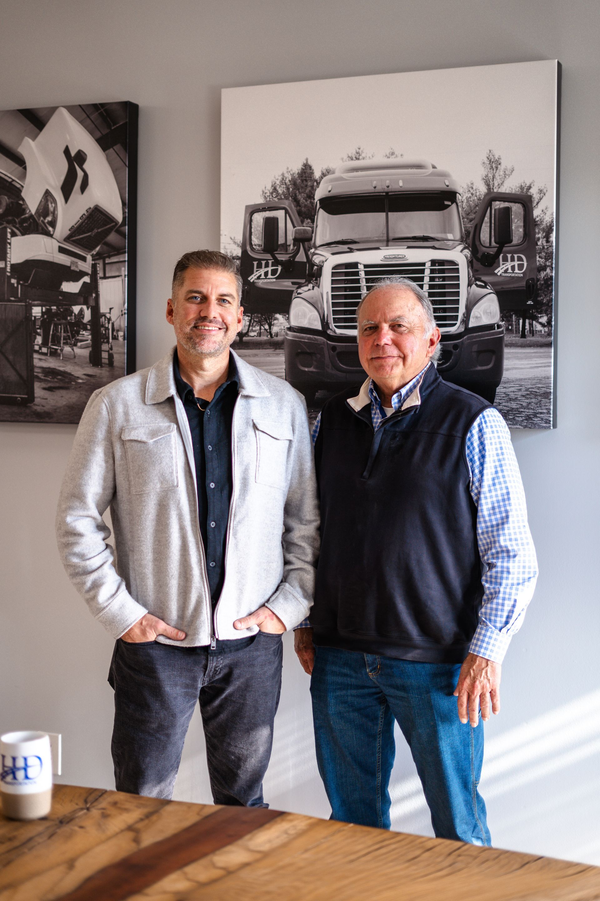 Two men stand by a wooden table, smiling. One wears a jacket, the other a vest. Artwork of a truck hangs on the wall.