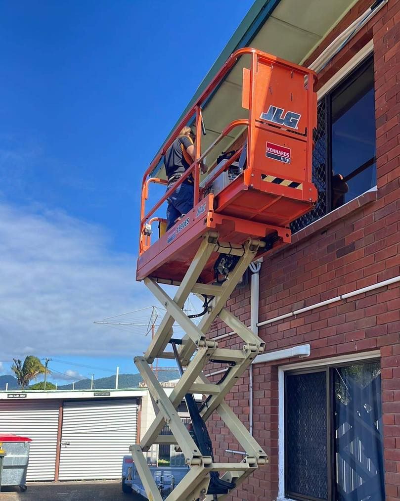 A Man Is Standing on A Scissor Lift in Front of A Brick Building — Recharged Air & Electrical in Gordonvale, QLD