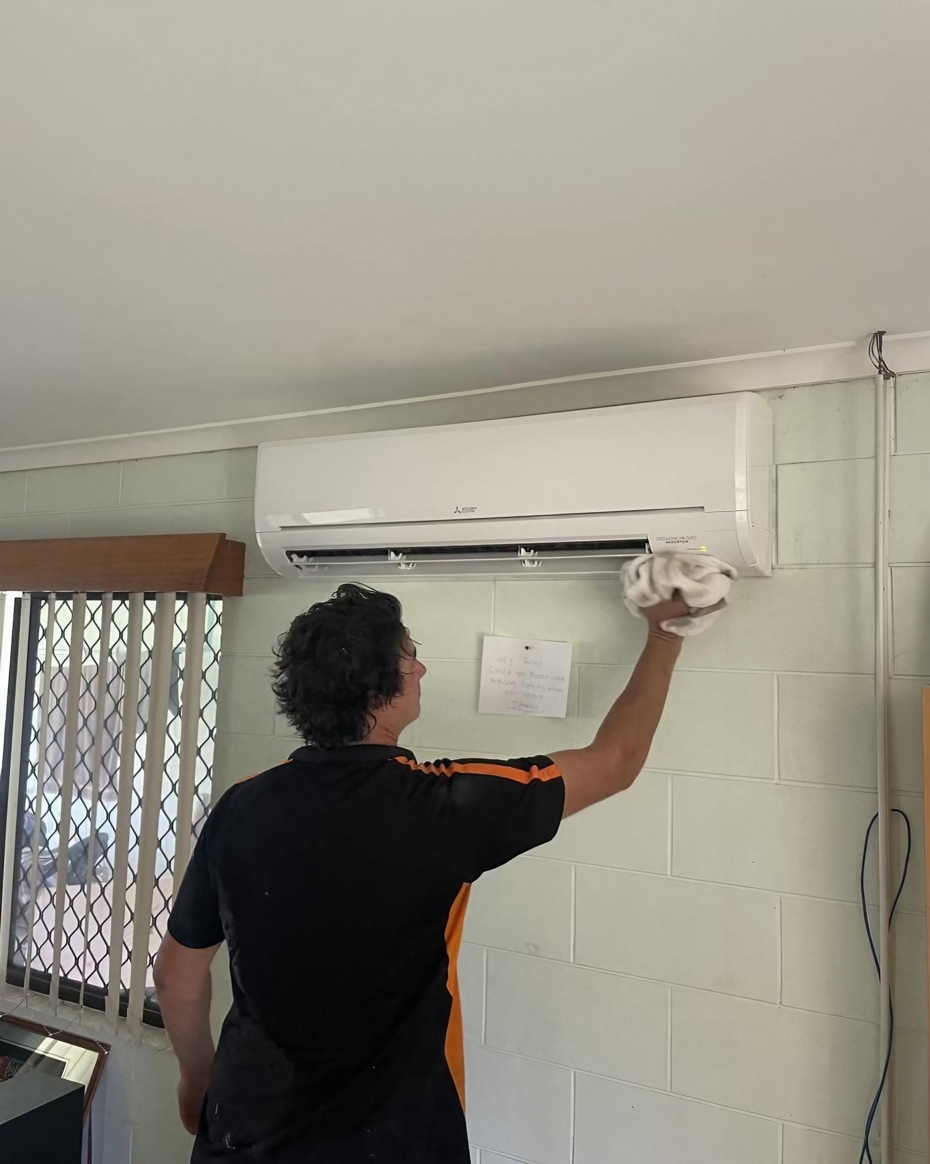Man cleaning air conditioner with a white cloth, against a white brick wall.