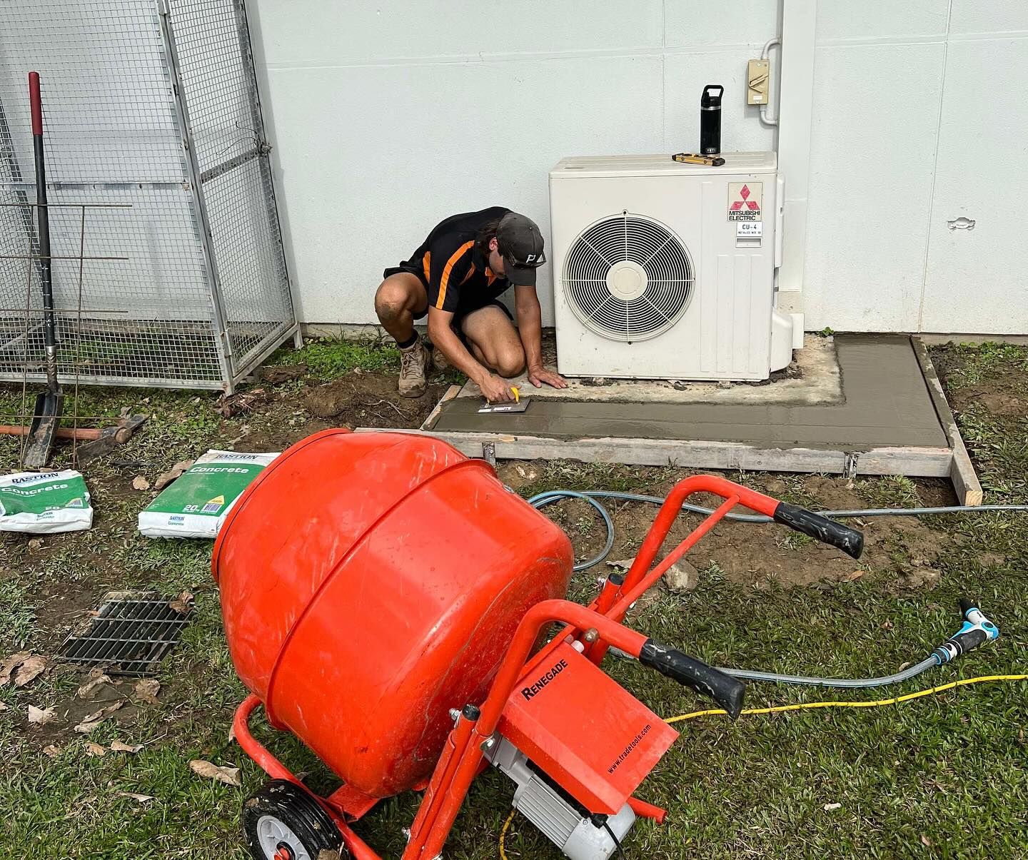 A Person Trowels Cement Around an Ac Unit on a Concrete Slab, With a Cement Mixer Nearby — Recharged Air & Electrical in Cairns, QLD