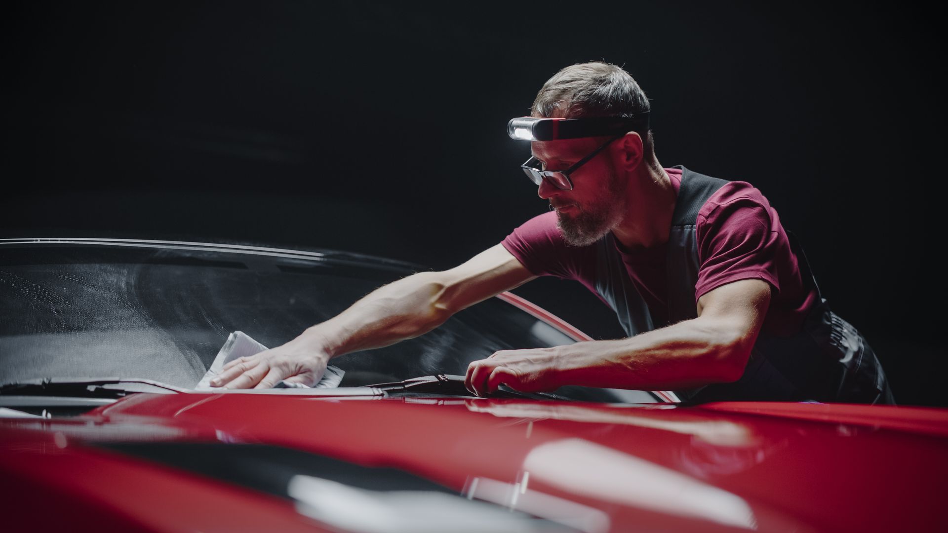 A man in a red shirt and headlamp cleans a red car windshield with a white cloth in a dark garage.