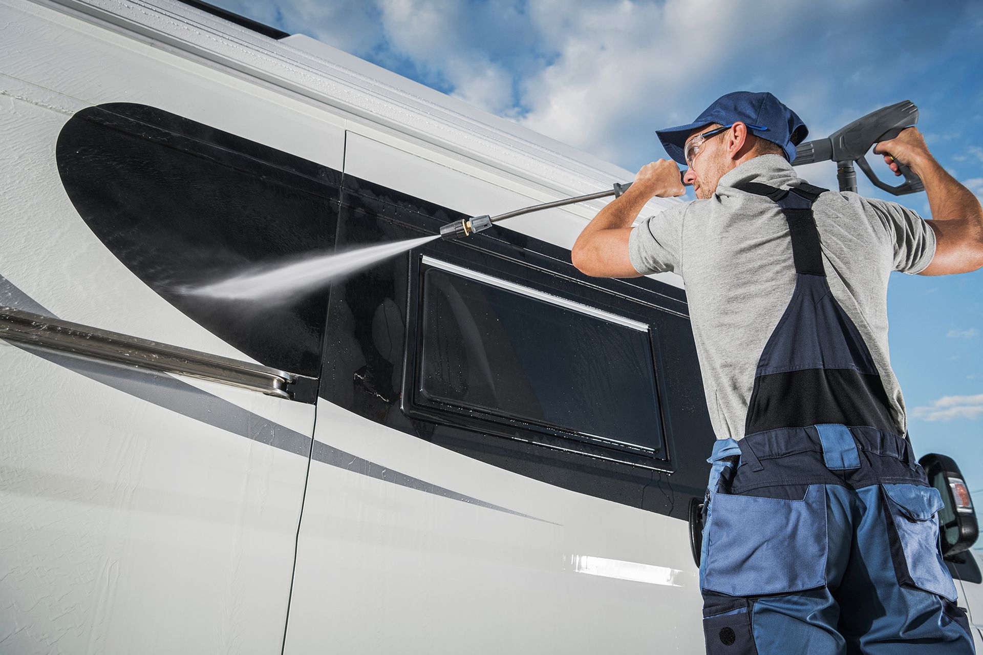 Man power washing a white aircraft with a pressure washer; blue sky background.