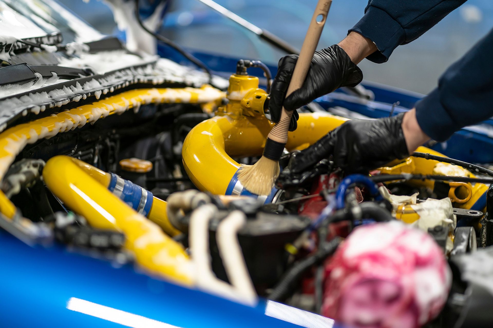 Hands in black gloves cleaning a car engine with a brush.