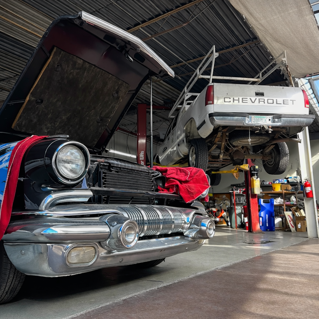 Classic car with open hood, a truck on a lift, both in a garage.