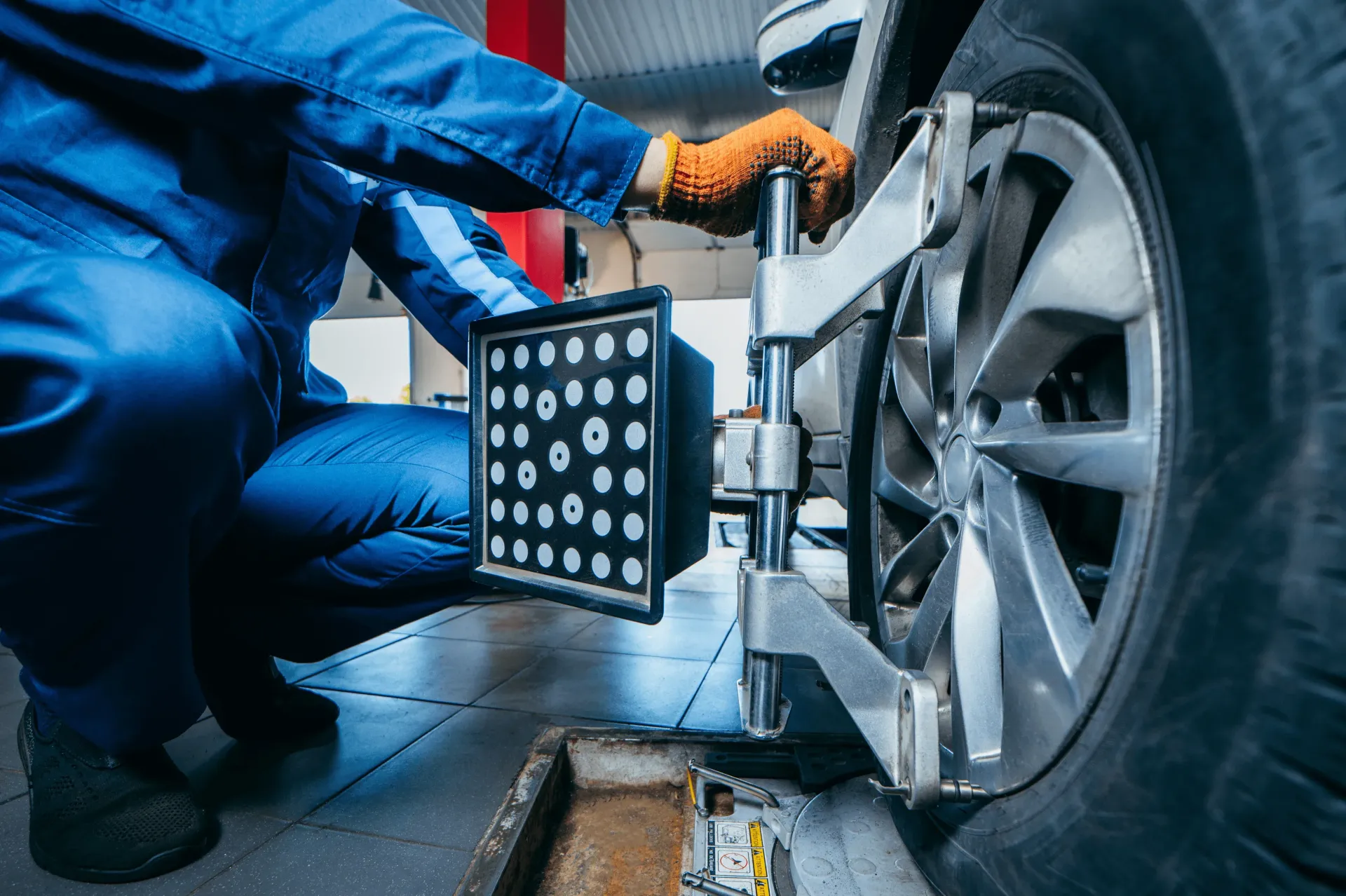 Mechanic in blue jumpsuit checks car wheel alignment with a calibration tool in a garage.