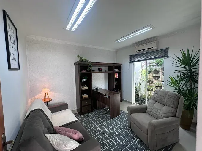 Escritório aconchegante com mesa, sofá, poltrona reclinável e plantas junto à janela.