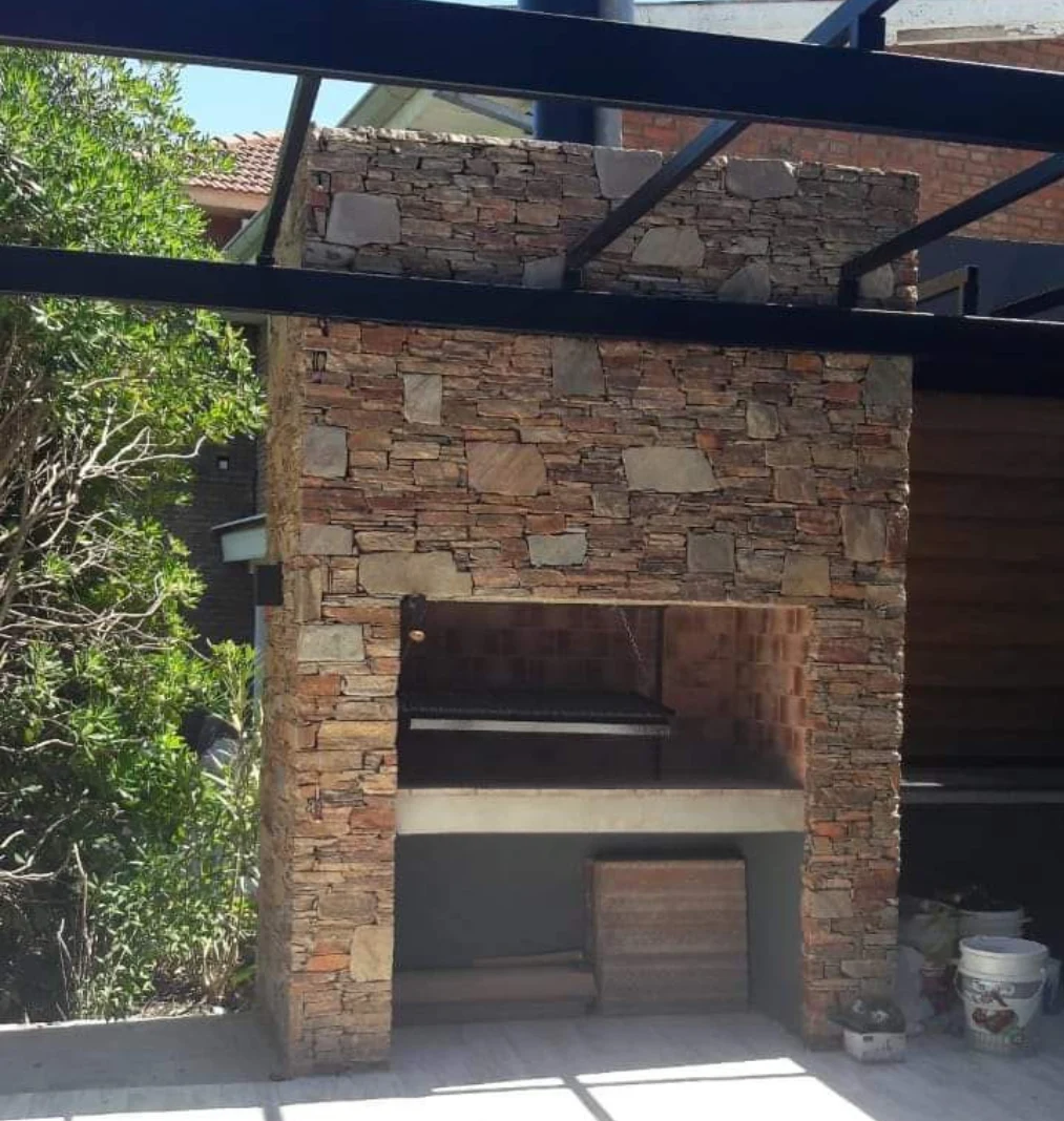 Chimenea de piedra al aire libre con techo de estructura metálica, junto a vegetación.