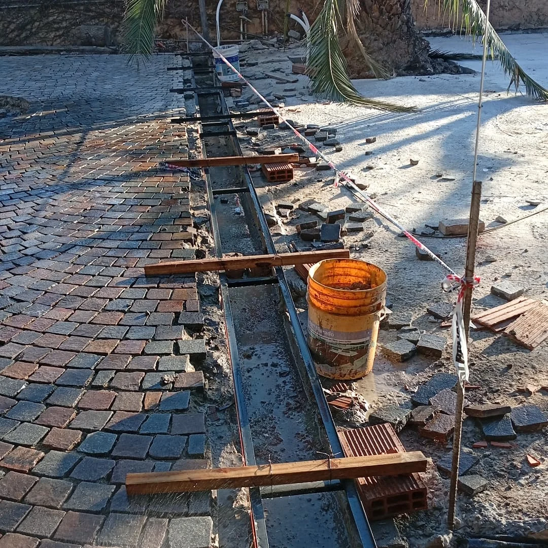 Obra con zanja y camino de ladrillos. Hay un cubo y soportes de madera.