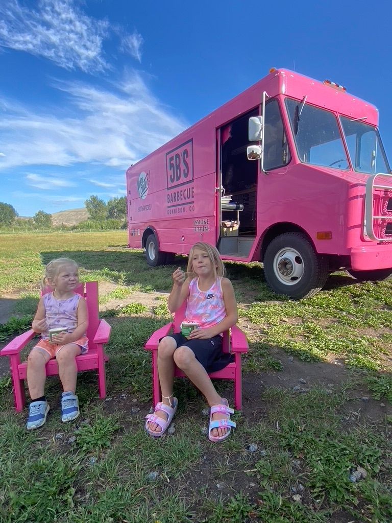 Two little girls are sitting in pink chairs in front of a pink food truck.