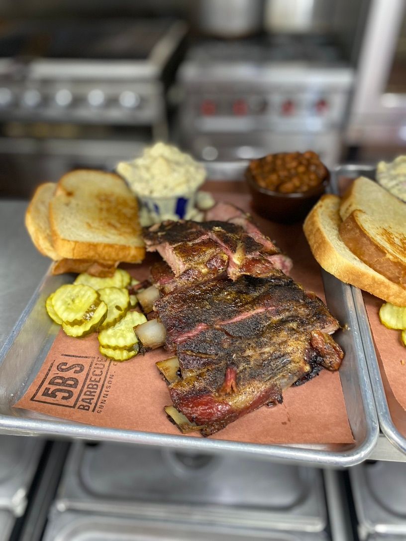 A tray of ribs , bread , pickles and beans on a table.