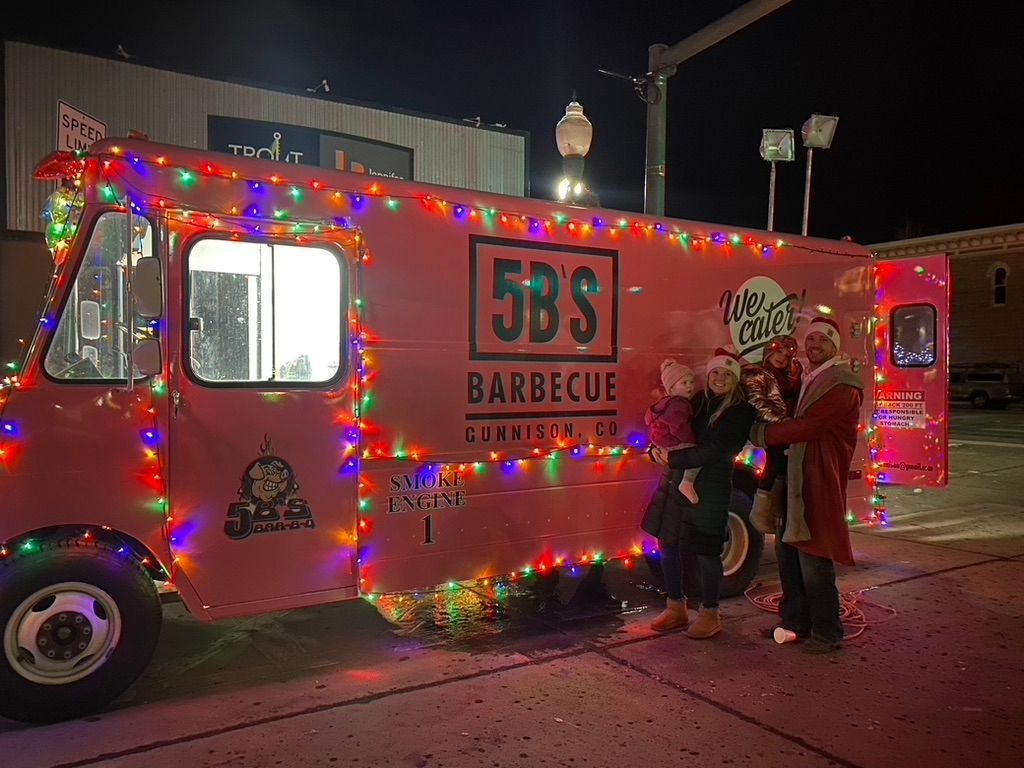 A group of people are standing in front of a pink food truck decorated with christmas lights.