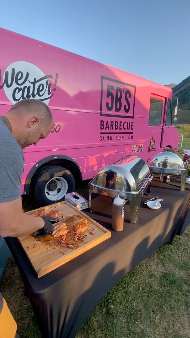 A man is cutting meat on a cutting board in front of a pink food truck.