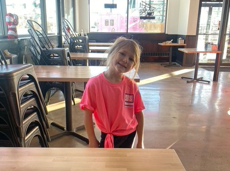 A little girl in a pink shirt is sitting at a table in a restaurant.