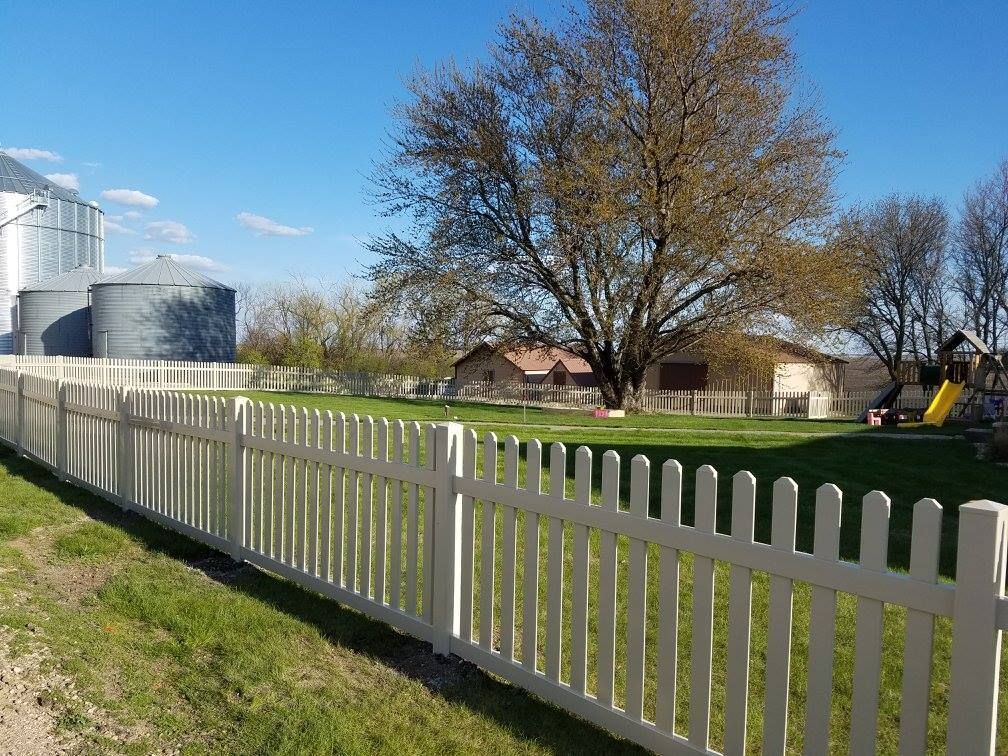 A white picket fence surrounds a grassy field with silos in the background.