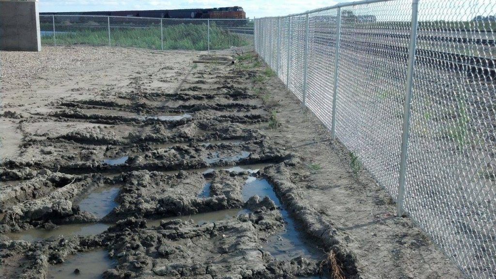 A chain link fence surrounds a muddy area with a train in the background.