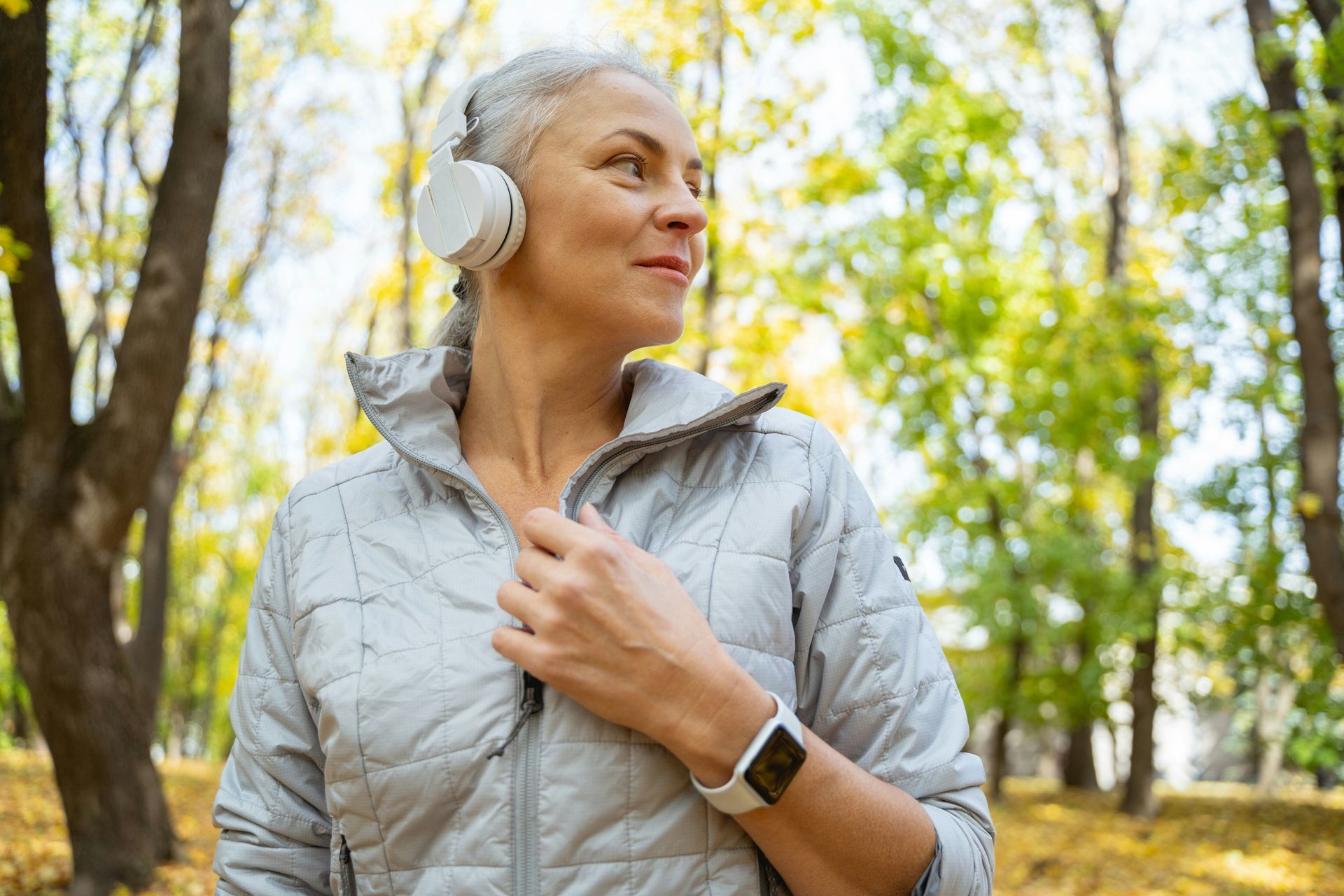 Woman in gray jacket, headphones, and watch, smiling in a park, surrounded by trees.